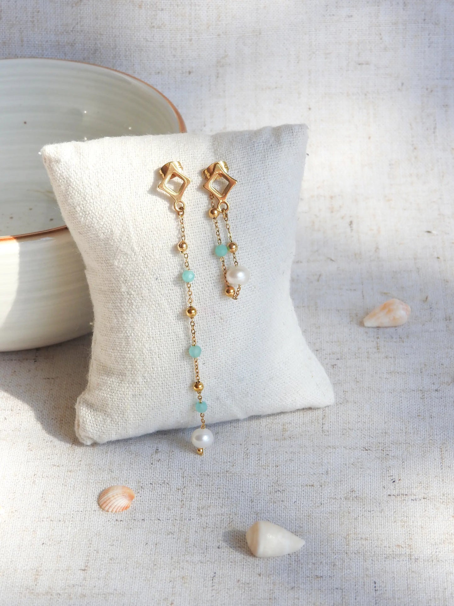 boucles d'oreilles clous losange en acier inoxydable doré. Chainettes pendantes asymétriques perlées en acier doré et perles de verre turquoise et perles d'eau douce blanche. Bijoux piqués sur un mini-coussin beige, contre un bol nu céramique, sur une table décorée de coquillages.