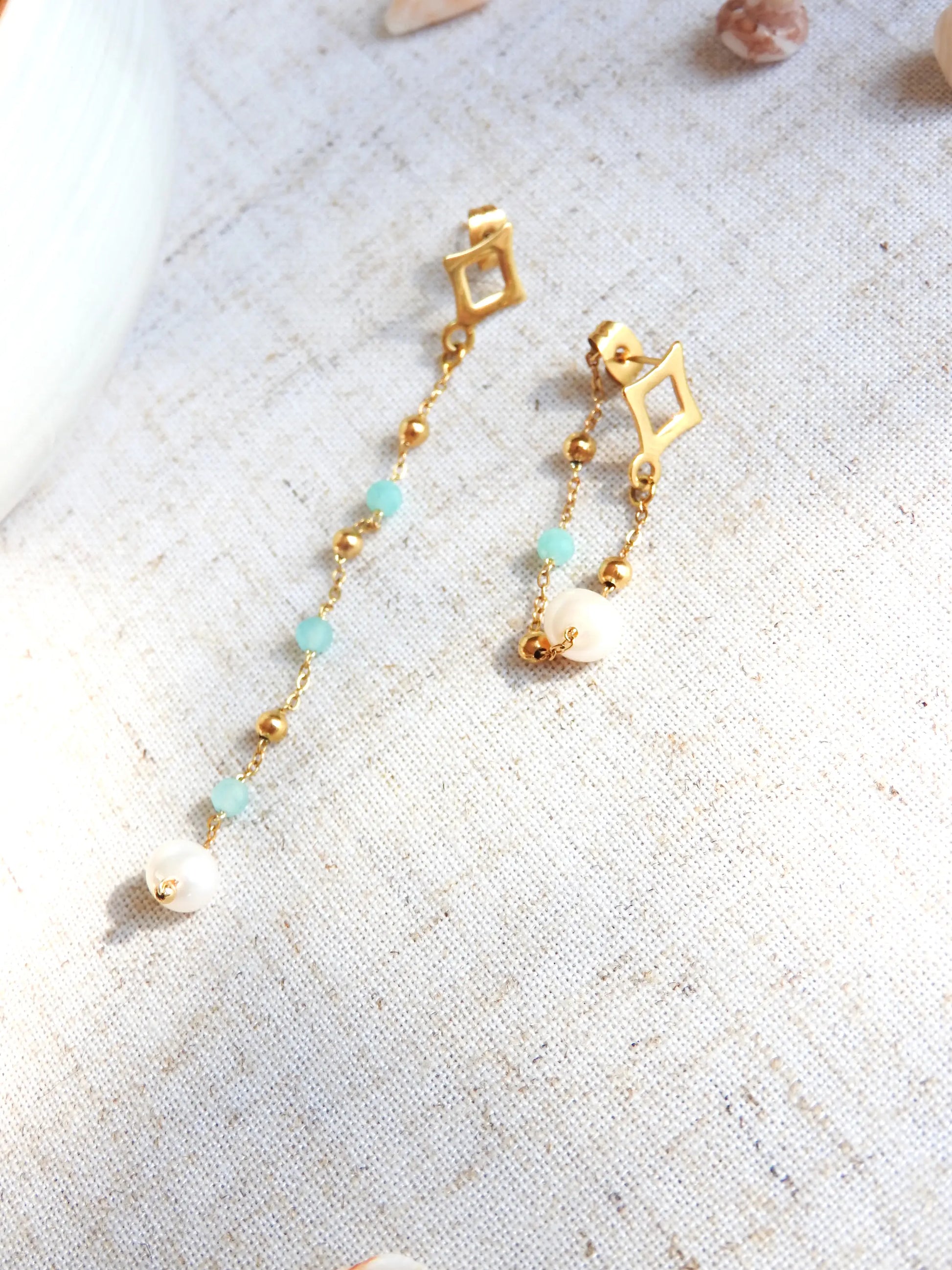 boucles-oreilles-pendantes-chaines-asymetriques-perles-acier-inoxydable-dore-et-perles-de-verre-turquoise-eau-douce-a-plat-maina
boucles d'oreilles clous losange en acier inoxydable doré. Chainettes pendantes asymétriques perlées en acier doré et perles de verre turquoise et perles d'eau douce blanche. Bijoux posés à plat sur une nappe beige.