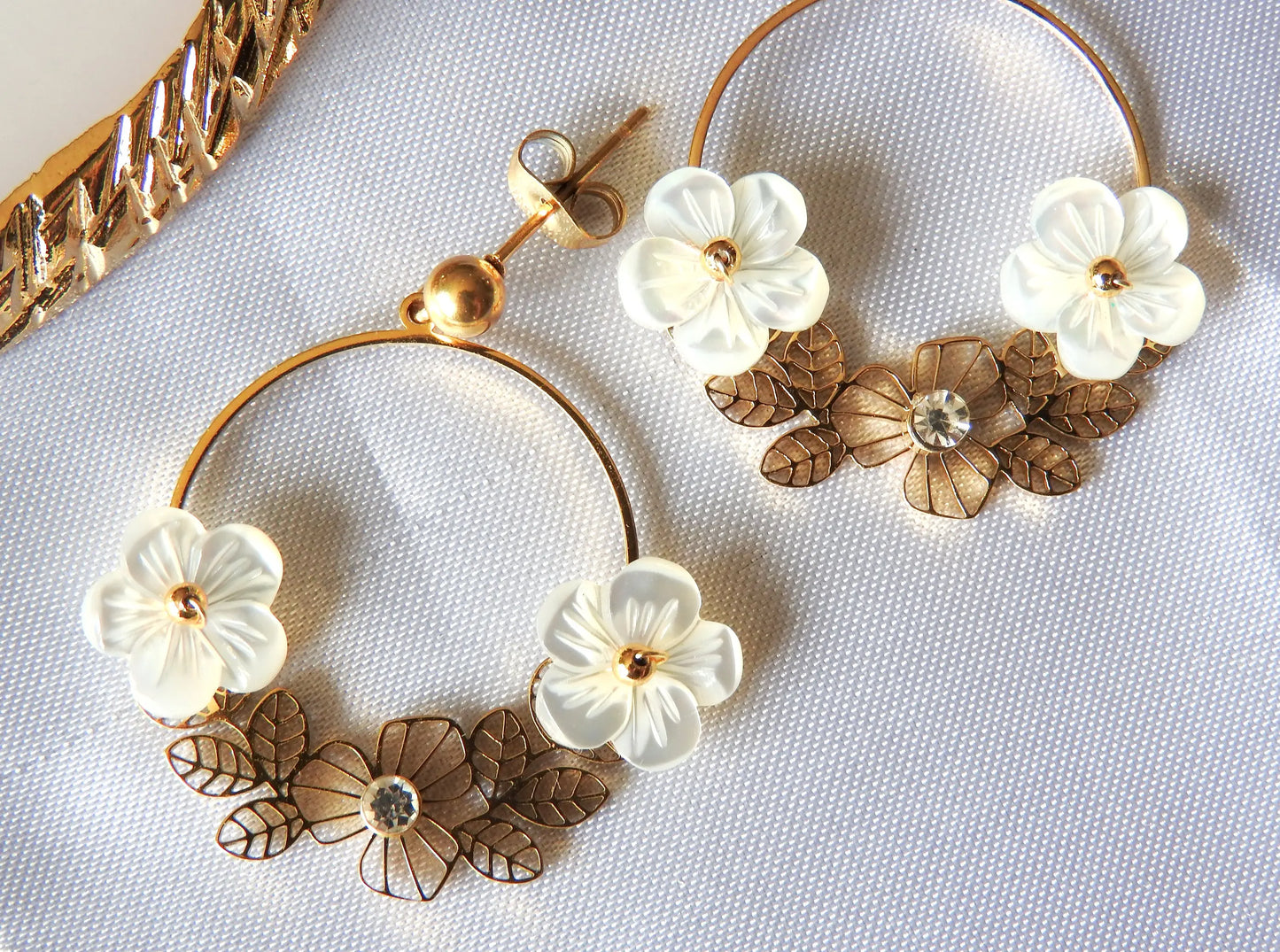 Boucles d'oreilles Jasmine de la marque de bijoux M'Joy Creation. Bijou posé à plat sur une nappe blanche. Boucles d'oreilles en créoles décorées de fleur blanche en nacre et strass.