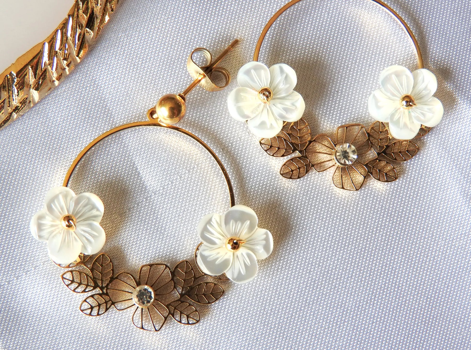 Boucles d'oreilles Jasmine de la marque de bijoux M'Joy Creation. Bijou posé à plat sur une nappe blanche. Boucles d'oreilles en créoles décorées de fleur blanche en nacre et strass.