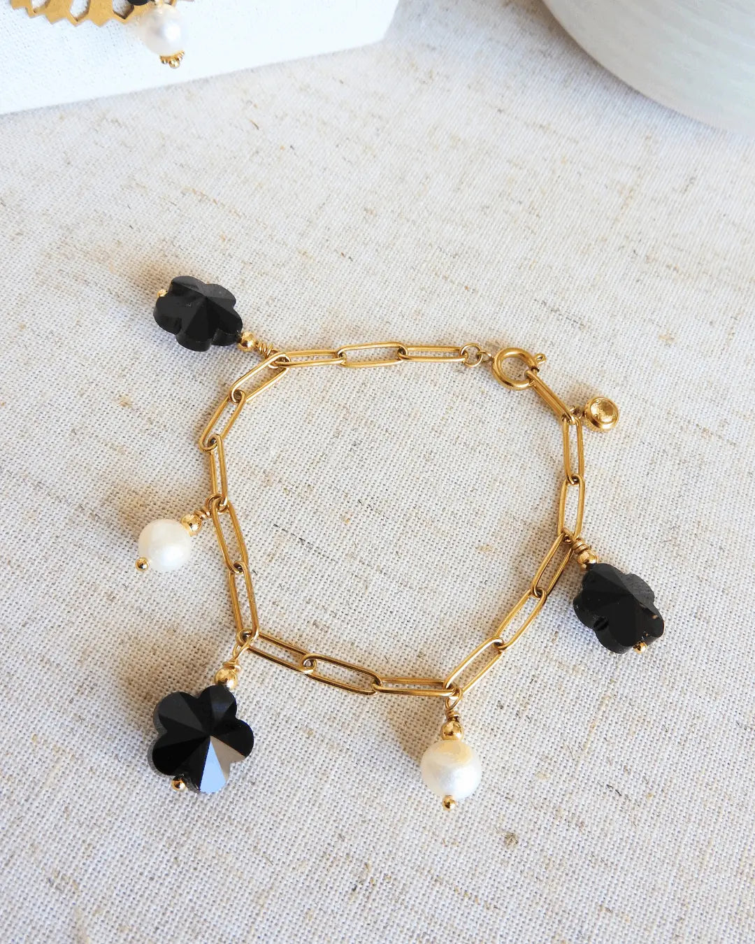 Bracelet posé à plat et fermé sur une nappe beige, bracelet chaine à maille rectangle en acier inoxydable doré, orné de breloques en perles d'eau douce blanche et perles fleurs en verre noire.