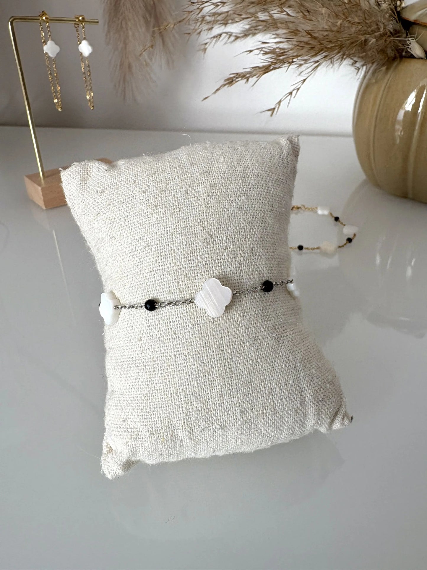 Bracelet chaîne en acier inoxydable argenté perlés de perles de verre noire ainsi que des perles trèfle en nacre blanche. Bijou présenté sur un mini-coussin beige en coton posé sur une table décorée de brindilles, un pot en céramique et des boucles d'oreilles pendantes.
