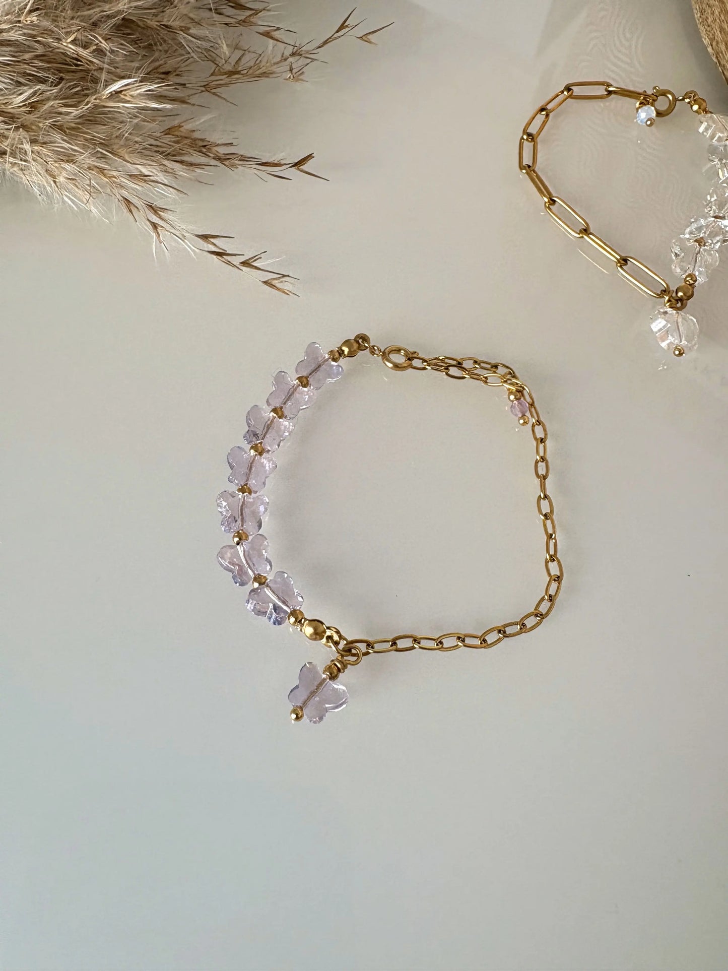 Bracelet mi-chaîne en acier inoxydable et perles de verre de forme papillon mauve, posé sur une table en verre trempée et décorée d'une brindille.