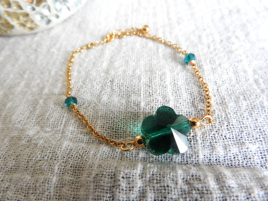 Bracelet chaîne en acier inoxydable doré, orné d'une perle de verre en forme de fleur couleur émeraude et de deux perles de chaque côté du bracelet du même couleur. Bijou posé sur une nappe blanche en gaze de coton.