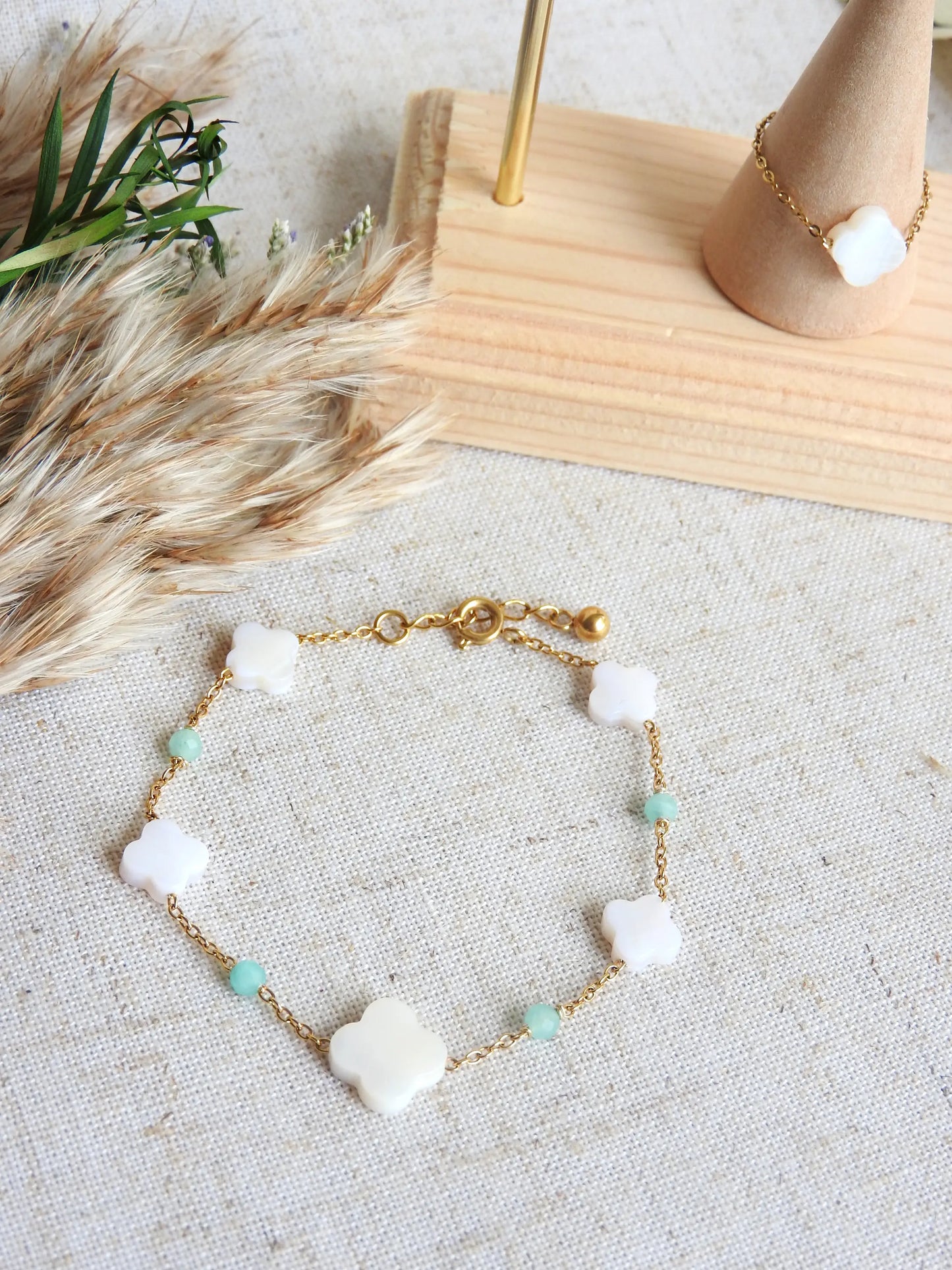 bracelet chaîne perlées en verre turquoise et perles trèfle en nacre blanche. Bijou en acier inoxydable doré posé à plat sur une nappe beige décorée d'une bague chaînette avec une perle trèfle en nacre blanche posée sur un cône.