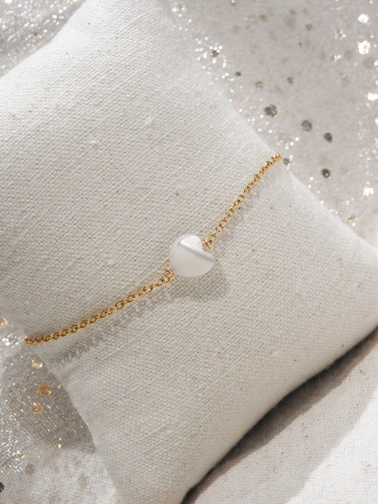 Bracelet chaîne fine en acier inoxydable doré orné d'une perle coeur en nacre blanche. Bijou posé sur un mini-coussin beige en coton.