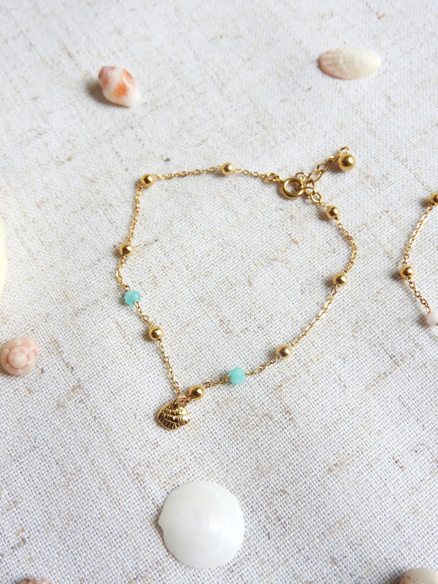 bracelet chaîne fine maille en acier inoxydable doré posé sur une nappe beige. Bracelet à chaine perlée en acier doré et perles de verre turquoise, orné d'une breloque coquillage.