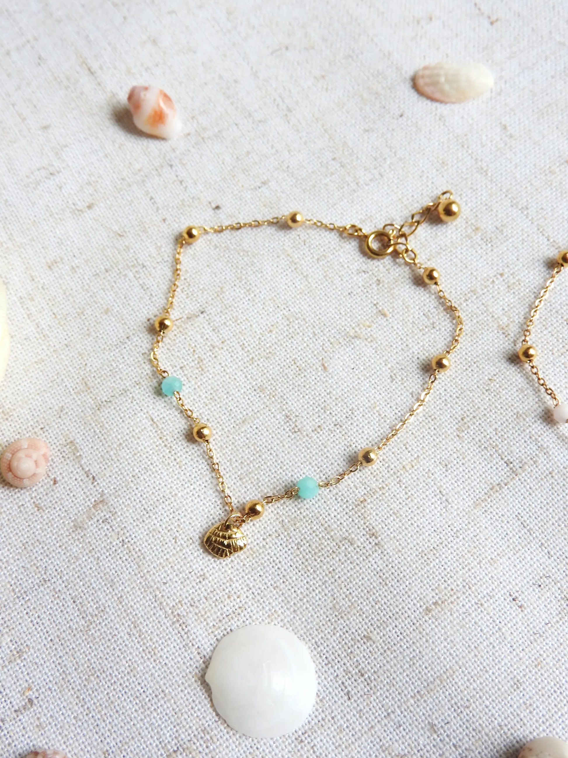 bracelet chaîne fine maille en acier inoxydable doré posé sur une nappe beige. Bracelet à chaine perlée en acier doré et perles de verre turquoise, orné d'une breloque coquillage.