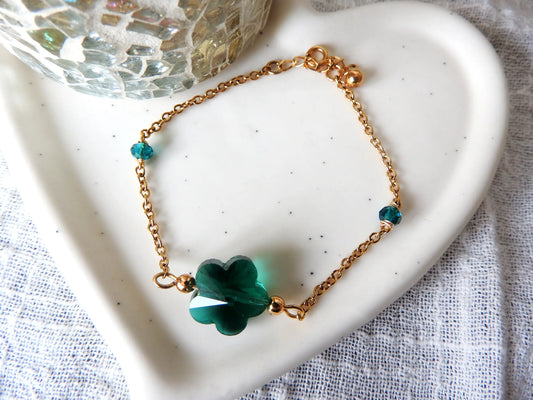 Bracelet chaîne en acier inoxydable doré, orné d'une perle de verre en forme de fleur couleur émeraude et de deux perles de chaque côté du bracelet du même couleur. Bijou posé sur une coupelle en céramique blanche.