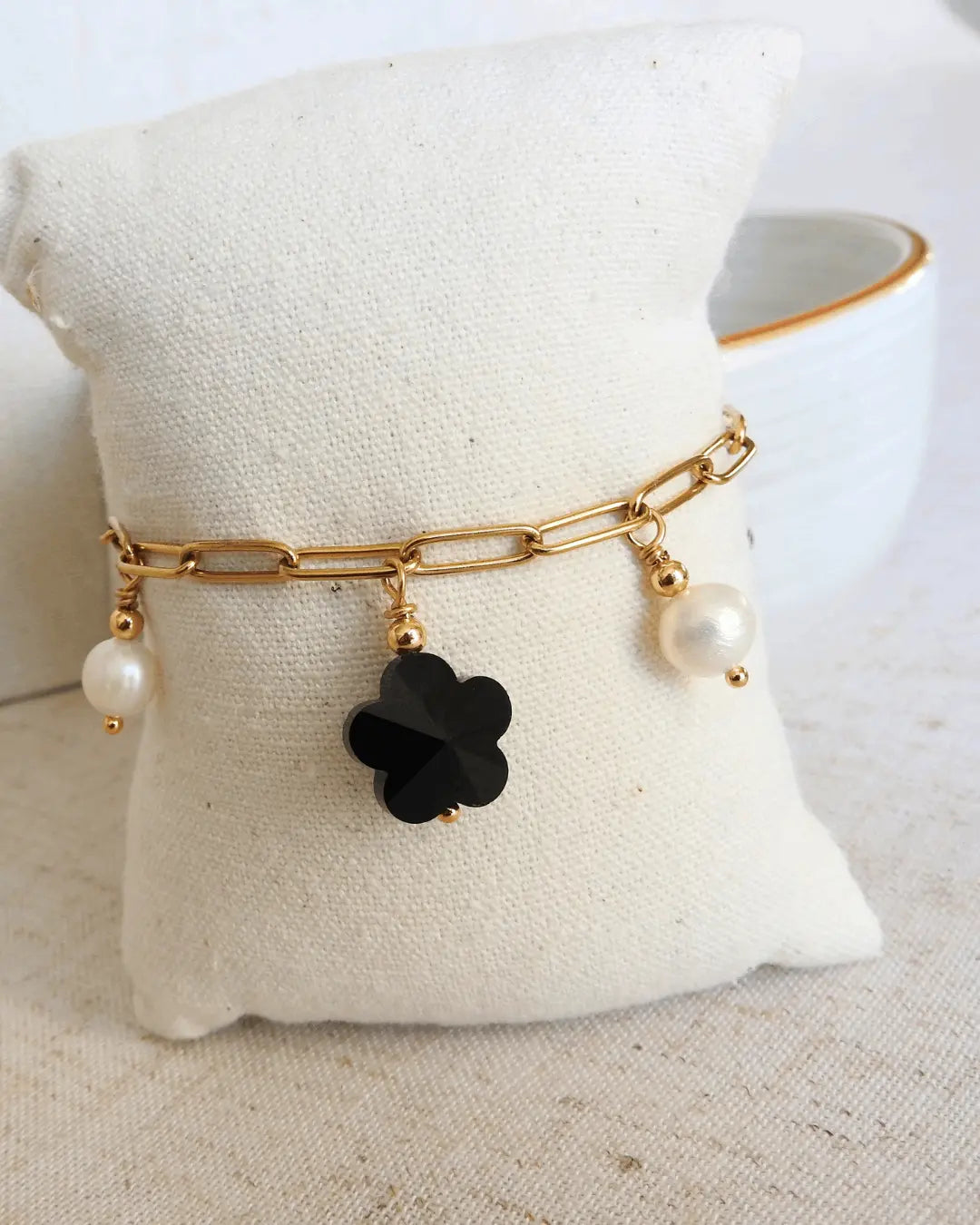 Bracelet présenté sur un mini-coussin beige, composé d'un bracelet chaine à mailles rectangle en acier inoxydable doré, orné de breloques en perles d'eau douce blanche et perles fleurs en verre noire.