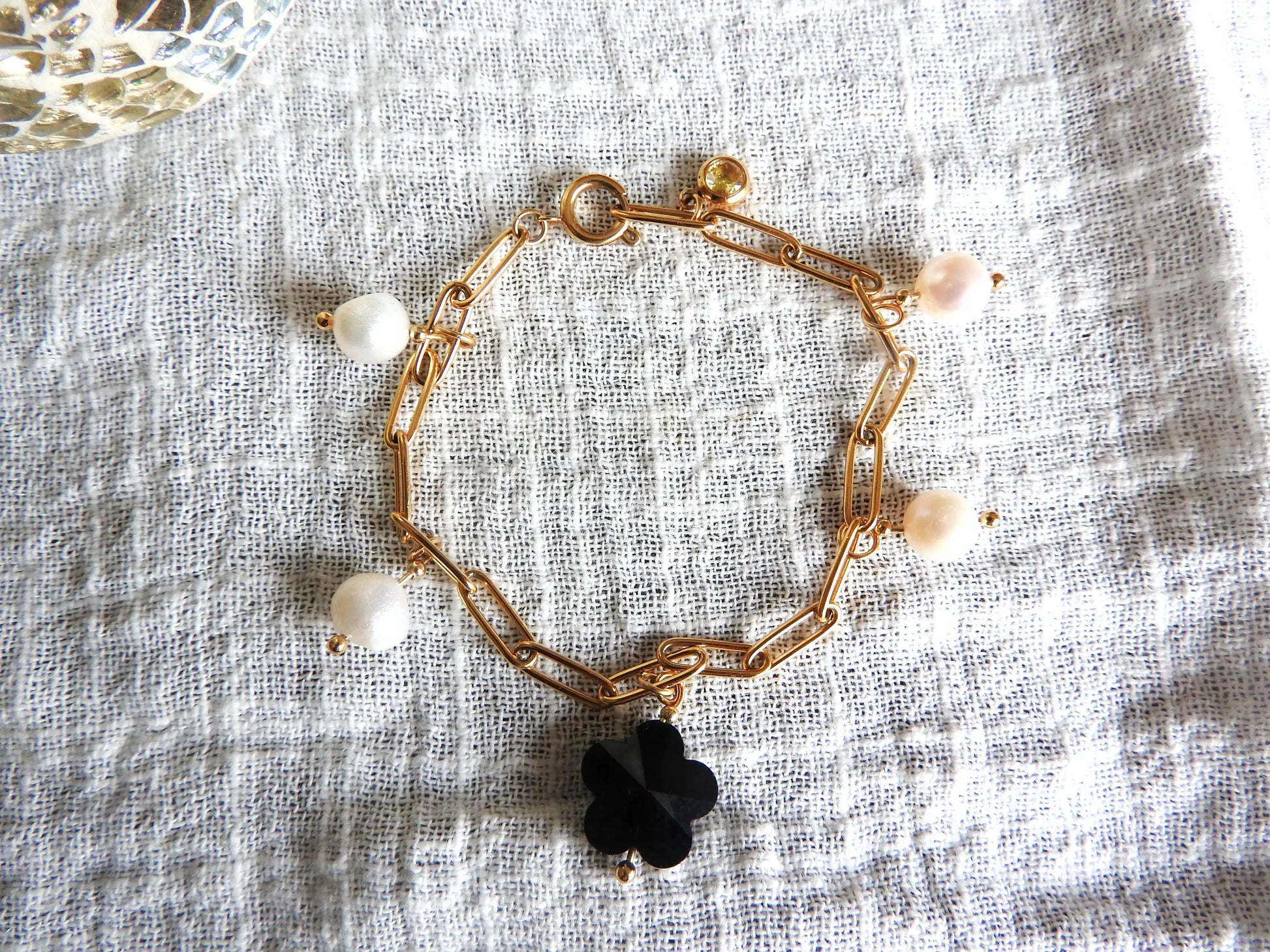 Bracelet chaine maille rectangle à breloques en acier inoxydable doré. Bijoux à breloques en perles d'eau douce blanche, perle en forme de fleur noire en verre et une pierre de naissance topaze. Bijou posé sur une nappe blanche en gaze de coton.