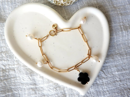 Bracelet chaine maille rectangle à breloques en acier inoxydable doré. Bijoux à breloques en perles d'eau douce blanche, perle en forme de fleur noire en verre et une pierre de naissance topaze. Bijou posé sur une coupelle en céramique blanche.