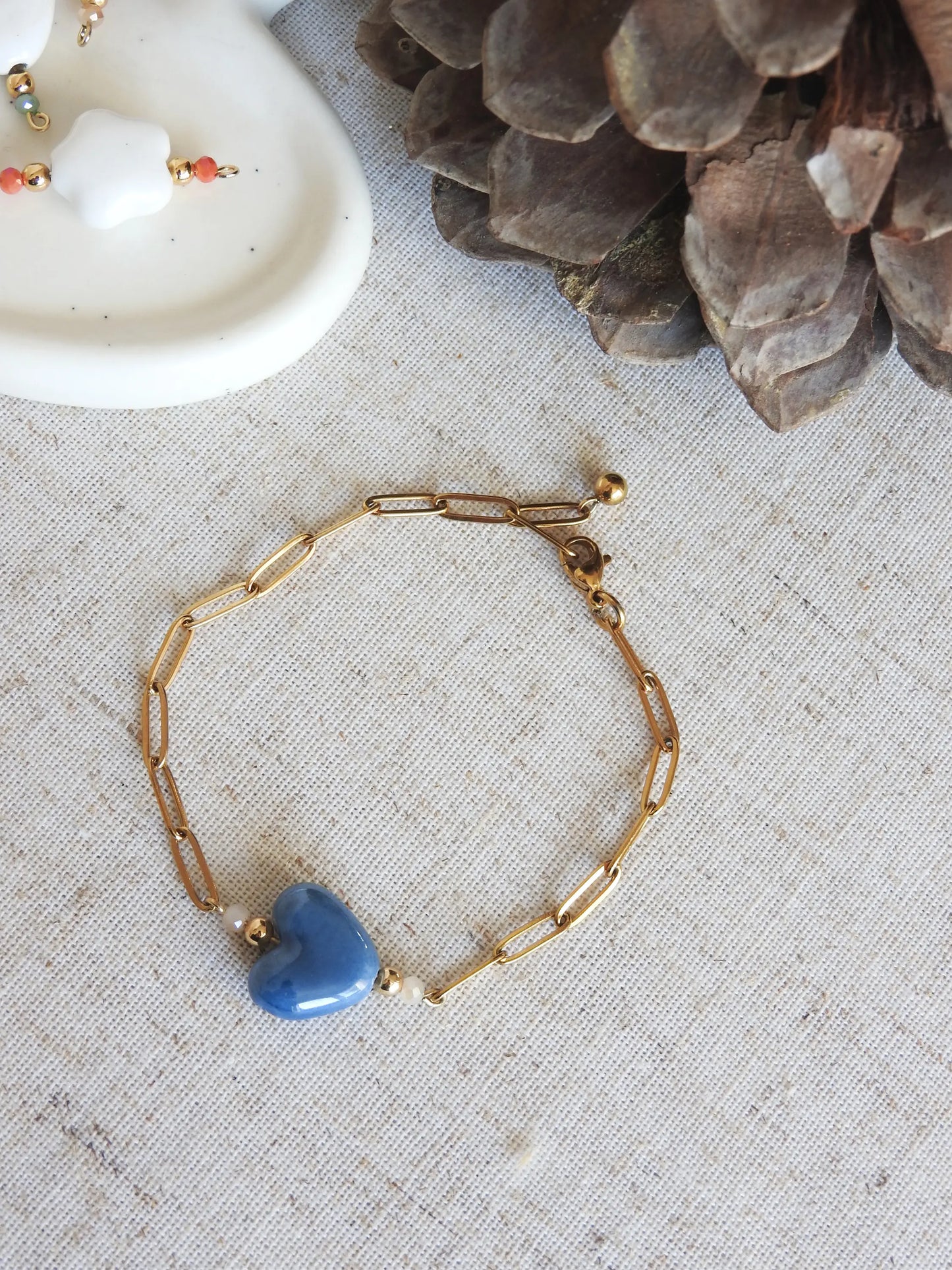 Bracelet chaîne mailles rectangle en acier inoxydable doré et orné de perles en verre et une perle fleur en céramique  bleu. Bijou posé à plat sur une nappe beige en coton.