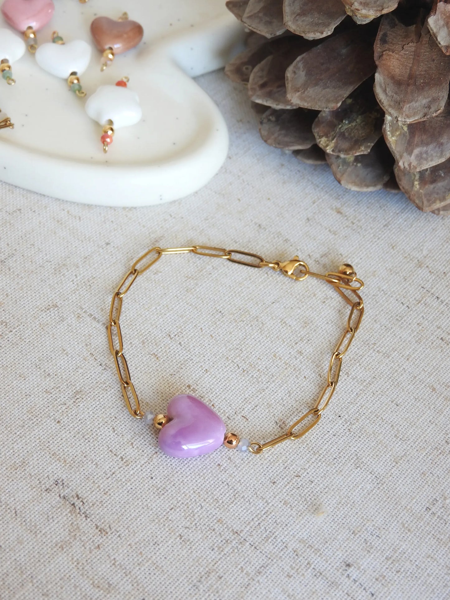 Bracelet chaîne mailles rectangle en acier inoxydable doré et orné de perles en verre et une perle fleur en céramique  mauve. Bijou posé à plat sur une nappe beige en coton.