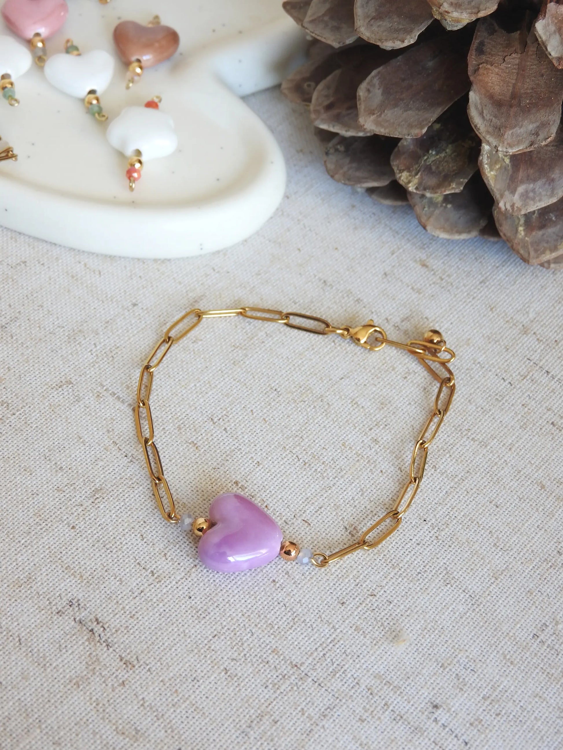 Bracelet chaîne mailles rectangle en acier inoxydable doré et orné de perles en verre et une perle fleur en céramique  mauve. Bijou posé à plat sur une nappe beige en coton.