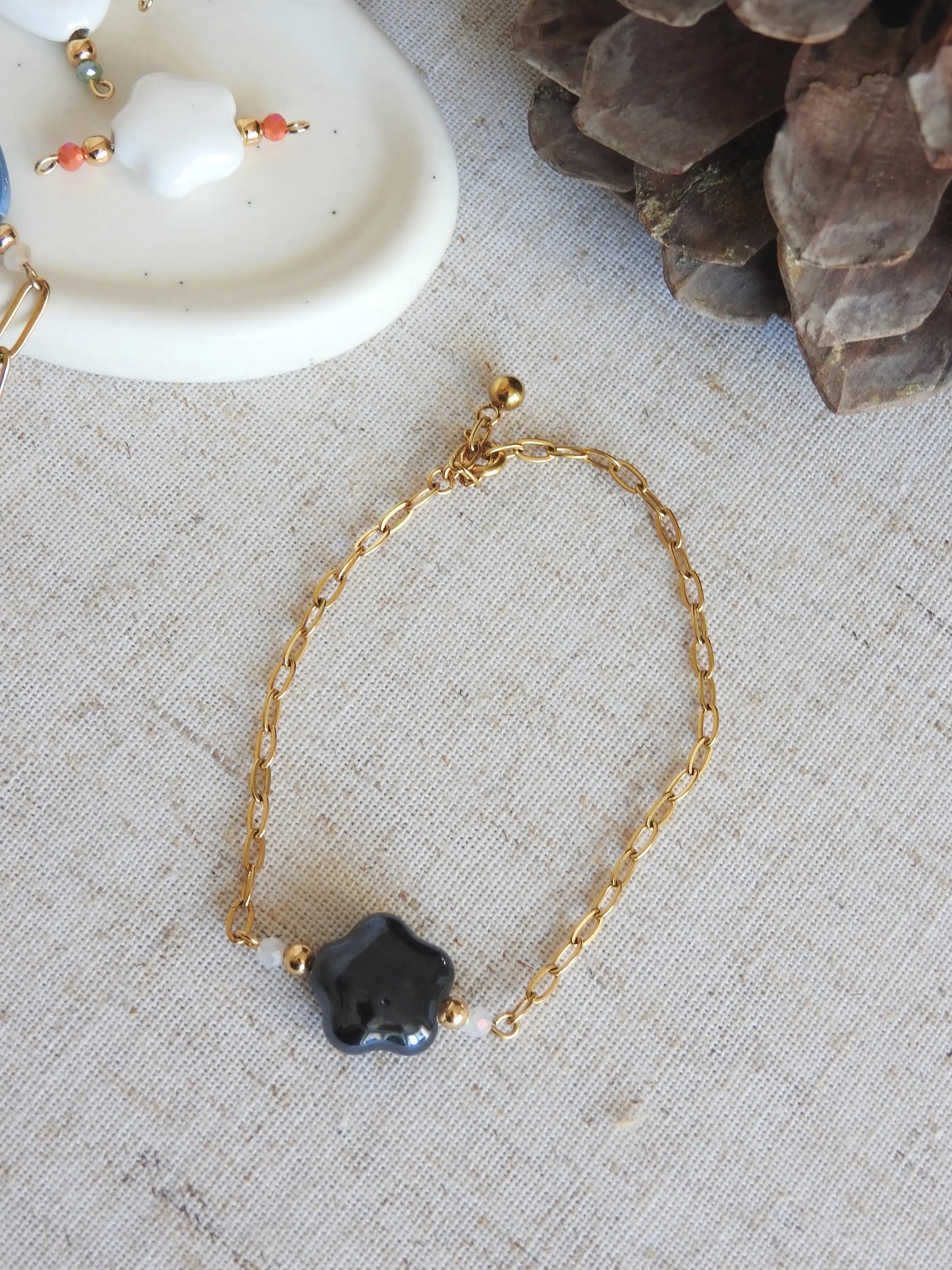 Bracelet chaîne mailles rectangle en acier inoxydable doré et orné de perles en verre et une perle fleur en céramique  noire. Bijou posé à plat sur une nappe beige en coton.