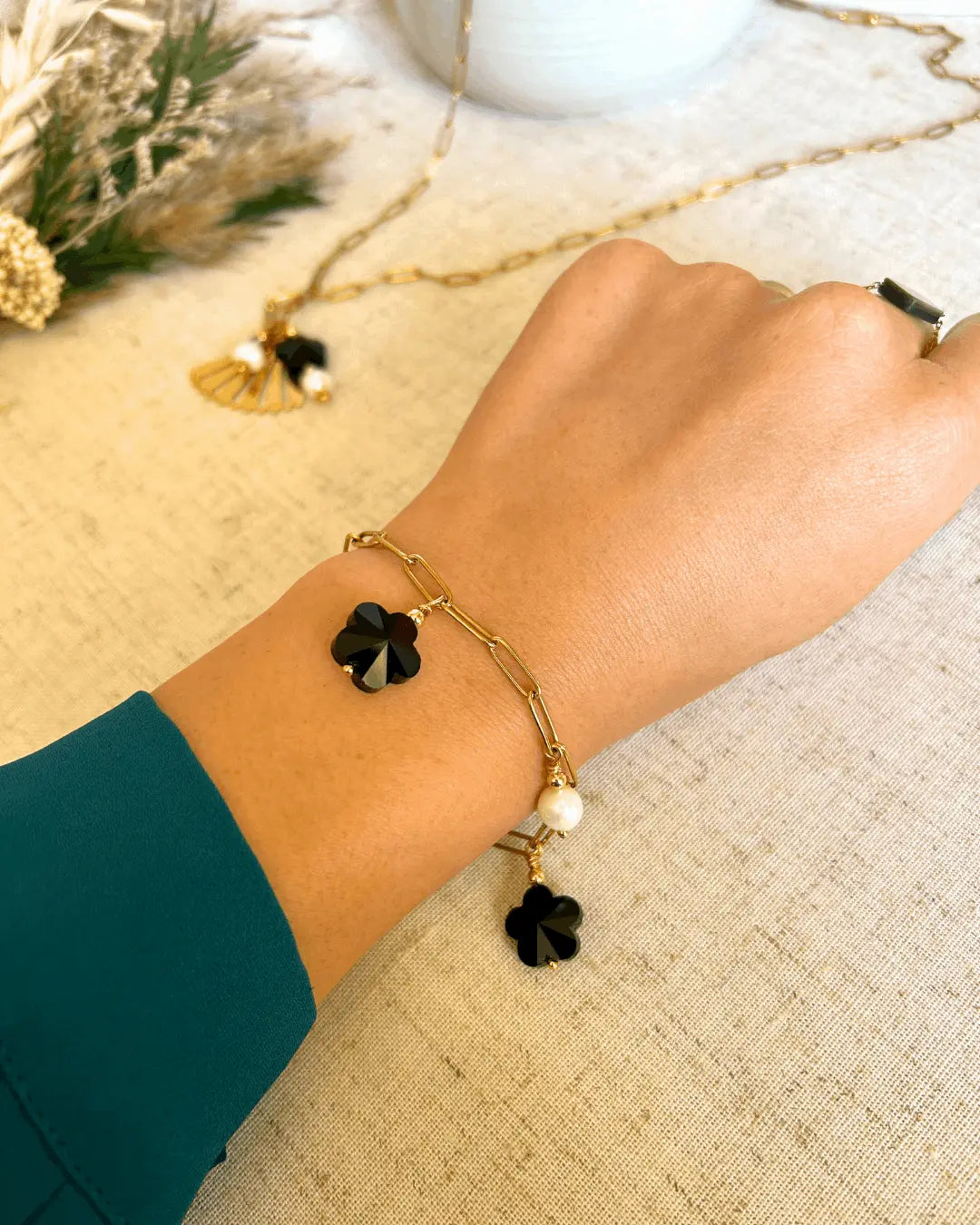 Bracelet posé à plat sur une nappe beige, composé d'un collier chaîne mailles rectangle en fond  En avant plan, un bracelet chaine maille rectangle en acier inoxydable doré, orné de breloques en perles d'eau douce blanche et perles fleurs en verre noire. Bijou porté au poignet posé sur la table.