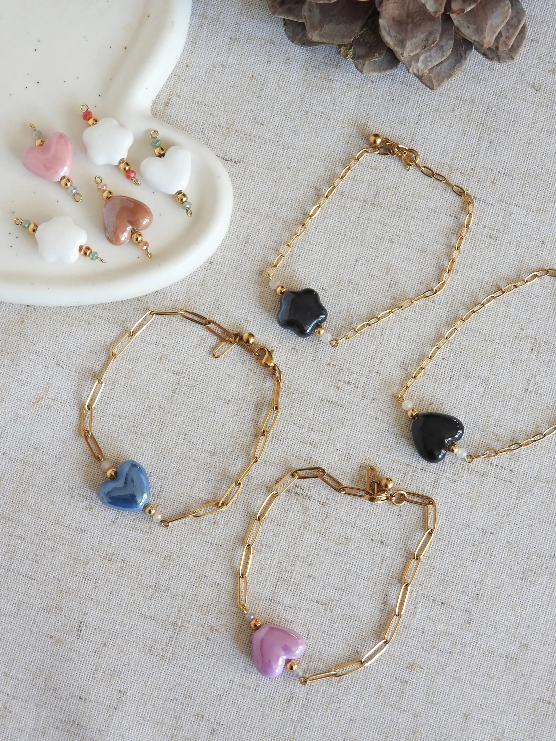 Bracelets chaîne mailles rectangle en acier inoxydable doré et ornées de perles de verre et perle coeur ou fleur en céramique de couleurs noire, bleu, rose, violet, marron et blanc. Bijoux disposés sur une nappe beige en coton.