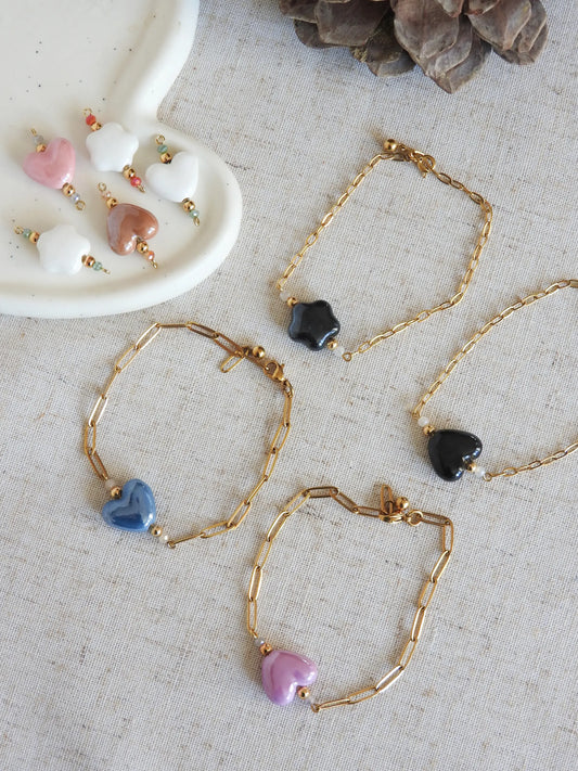 Bracelets chaîne mailles rectangle en acier inoxydable doré et ornées de perles de verre et perle coeur ou fleur en céramique de couleurs noire, bleu, rose, violet, marron et blanc. Bijoux disposés sur une nappe beige en coton.