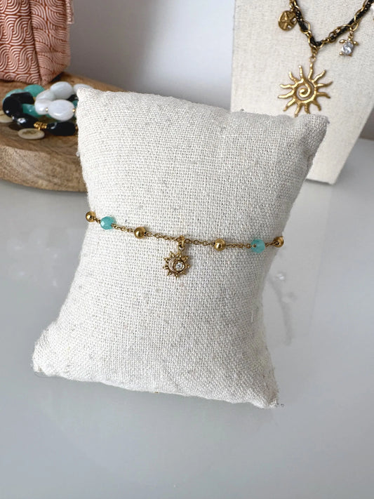 bracelet chaine perlées en acier inoxydable doré et perles turquoise amazonite, décoré d'une pampille soleil strass. Bijou présenté sur un mini-coussin en coton beige.