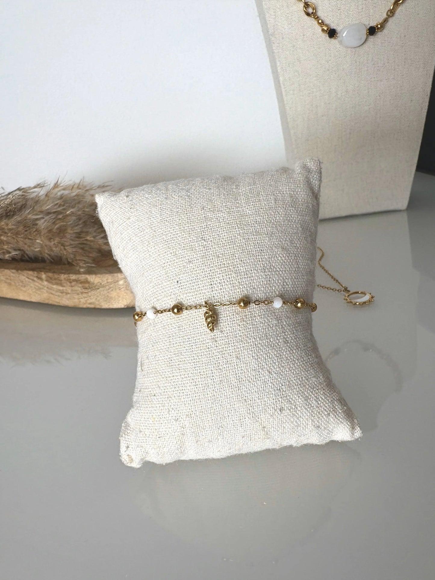 bracelet chaîne perlées en acier inoxydable doré et perles de verre blanc coquillage, décoré d'une pampille soleil strass. Bijou présenté sur un mini-coussin en coton beige, posé sur une table déocré d'un plateau en bmabou et buste présentoir et un collier chaine.