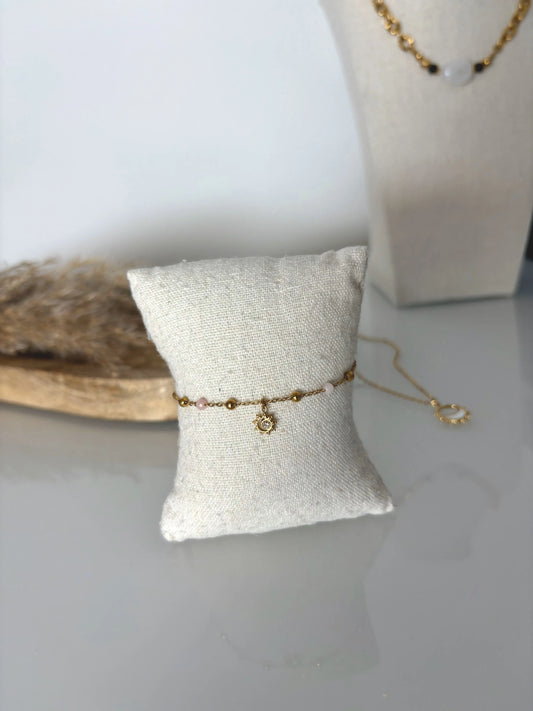 bracelet chaîne perlées en acier inoxydable doré et perles de verre rose pâle, décoré d'une pampille soleil strass. Bijou présenté sur un mini-coussin en coton beige, posé sur une table déocré d'un plateau en bmabou et buste présentoir et un collier chaine.