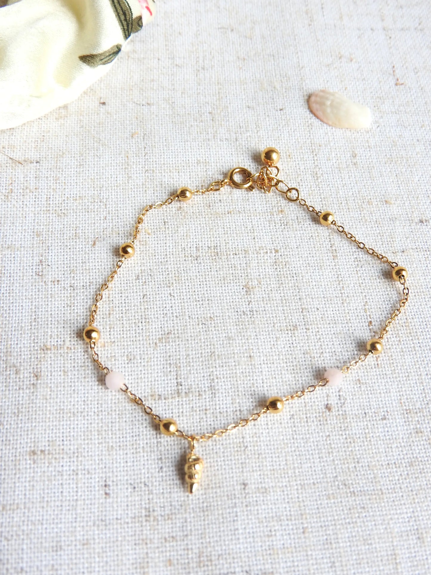 bracelet à chaine fine perlées en verre et acier inoxydable doré, orné d'une mini-breloque coquillage doré. Bijou posé sur une nappe beige.