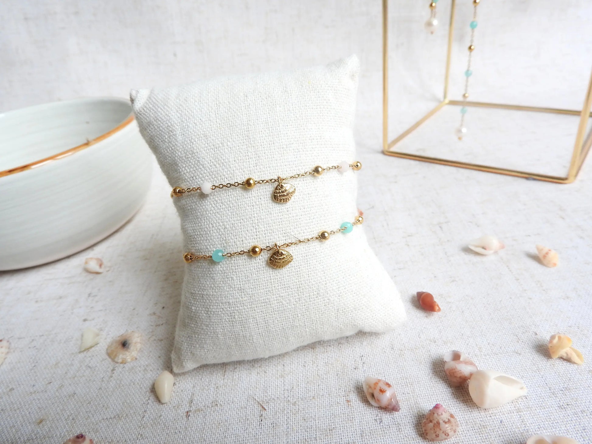 deux bracelets présentés sur un mini-coussin beige en coton. Bracelet chaîne perlées en verre et acier inoxydable doré, ornés d'une breloque coquillage en acier doré.