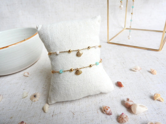 deux bracelets présentés sur un mini-coussin beige en coton. Bracelet chaîne perlées en verre et acier inoxydable doré, ornés d'une breloque coquillage en acier doré.