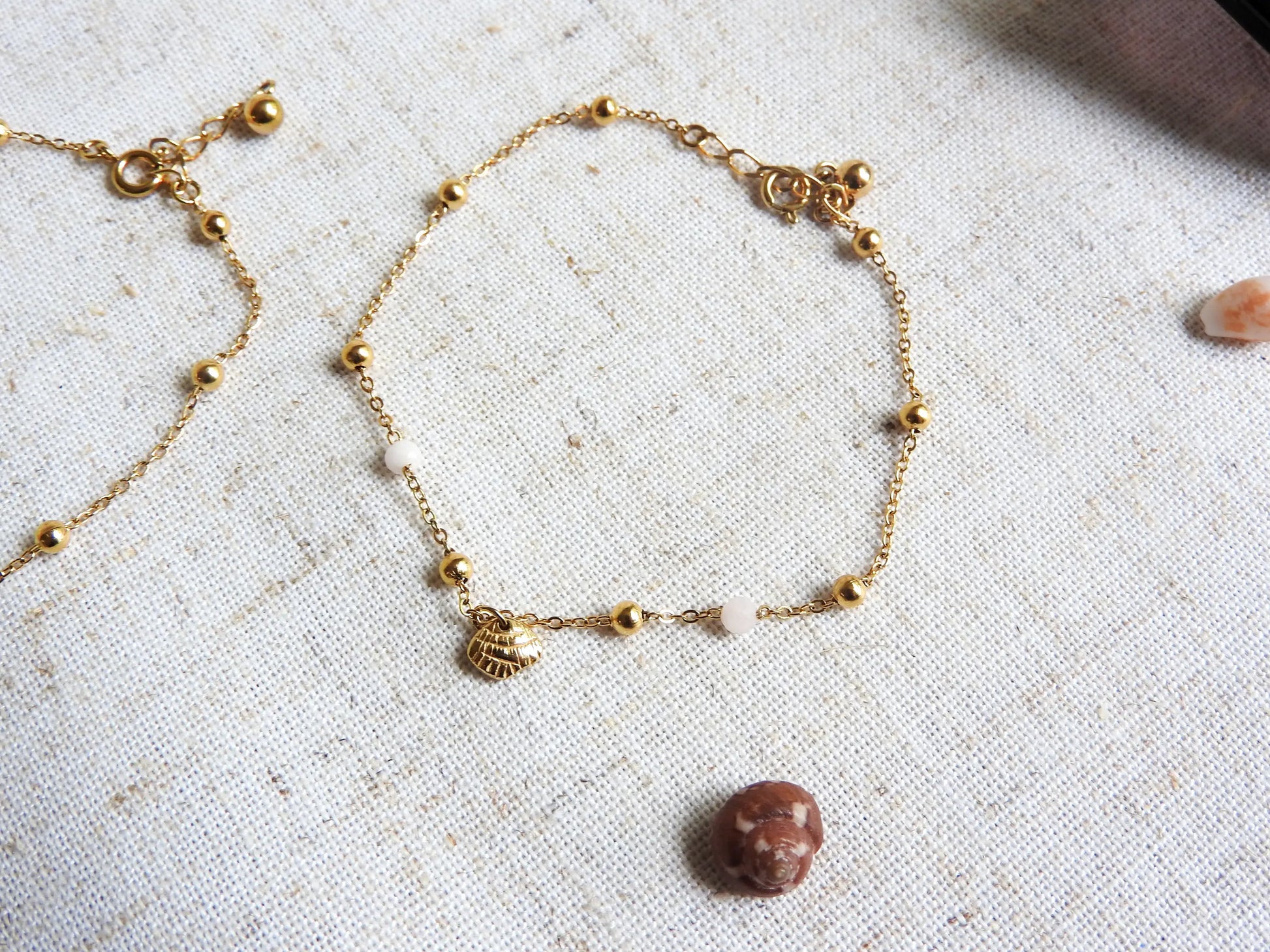 bracelet chaine perlée en verre et acier inoxydable doré posé à plat sur une nappe beige en coton. Bijou orné d'une breloque coquillage en acier doré.