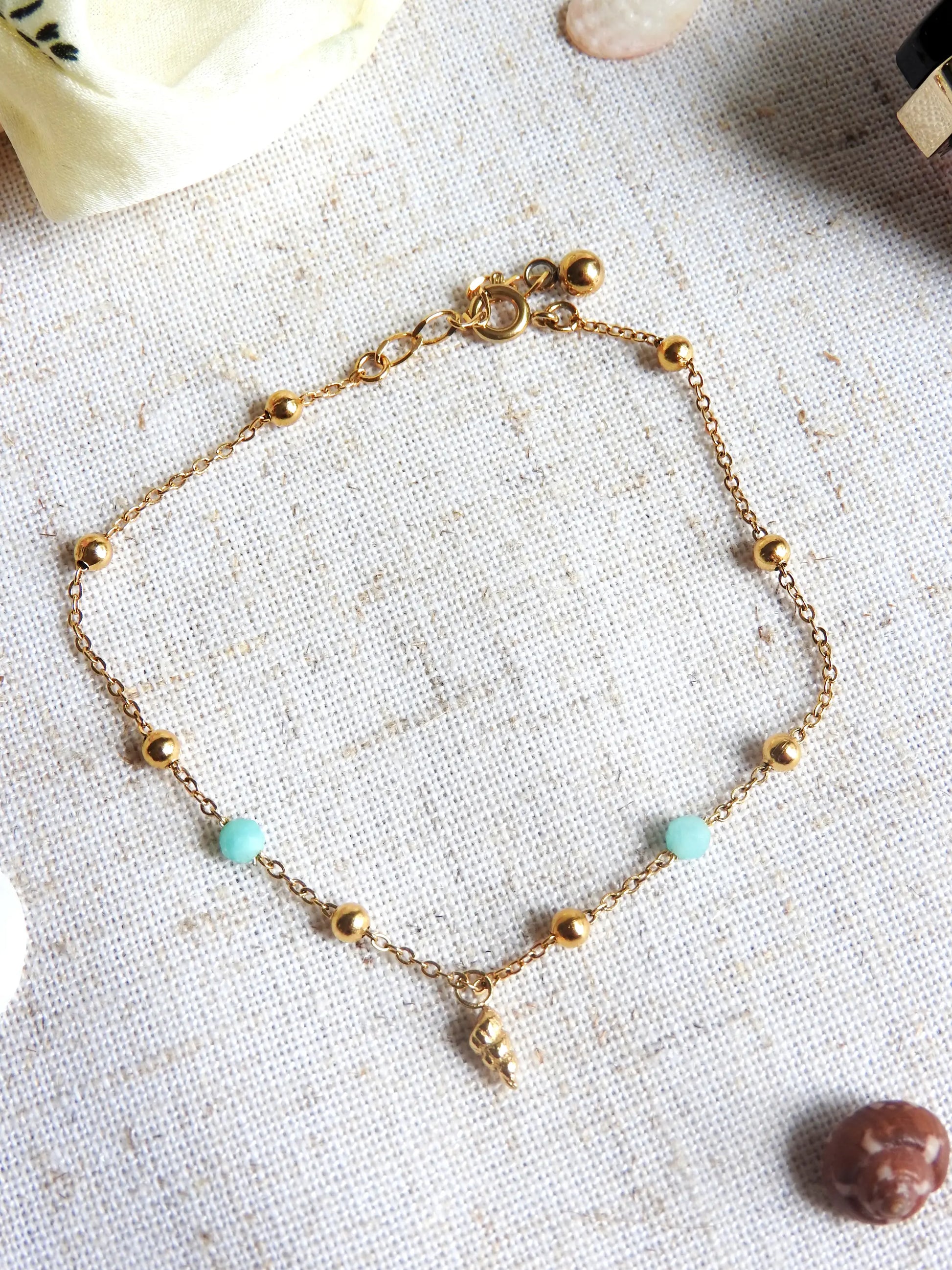 bracelet chaine fine maille perlée en verre et acier inoxydable doré, orné d'une mini-breloque coquillage doré. Bijou sur nappe beige en coton.