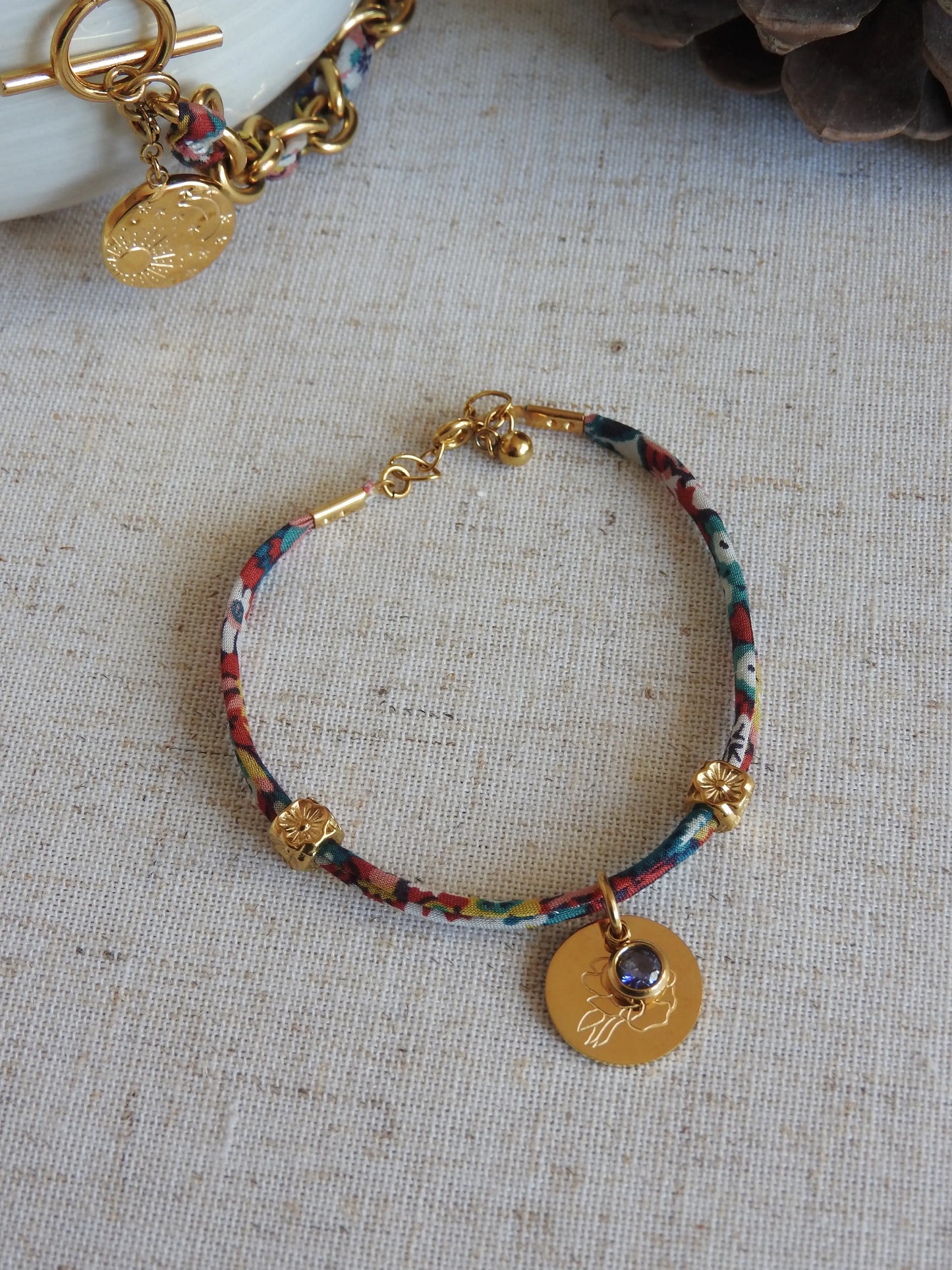 Bracelet cordon liberty Thorpe hill aux couleurs rose et blanc floral. Pendentif Fleur pivoine et pierre de naissance améthyste en acier inoxydable doré. Bijou présenté à plat sur une nappe beige en coton.