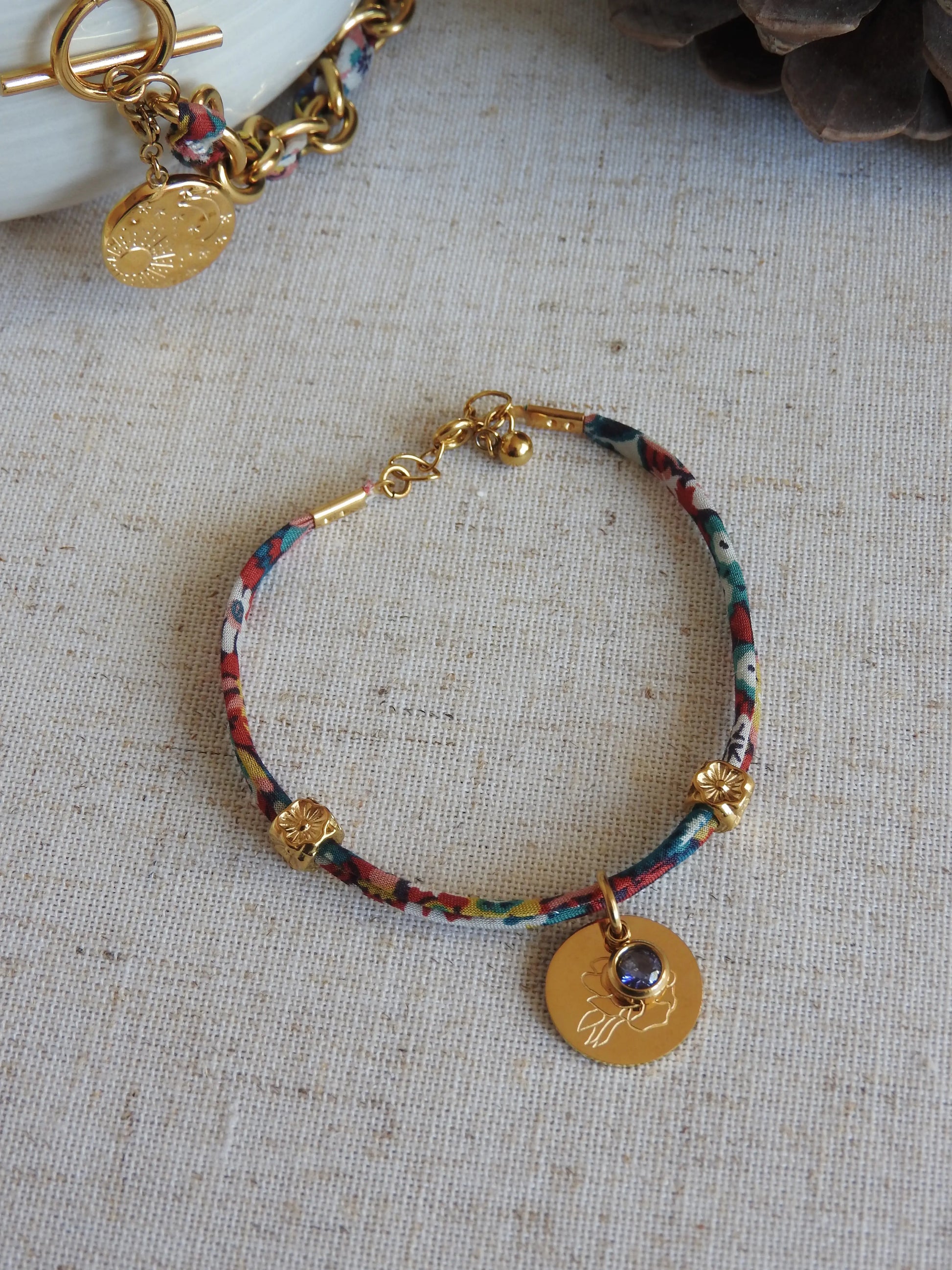 Bracelet cordon liberty Thorpe hill aux couleurs rose et blanc floral. Pendentif Fleur pivoine et pierre de naissance améthyste en acier inoxydable doré. Bijou présenté à plat sur une nappe beige en coton.
