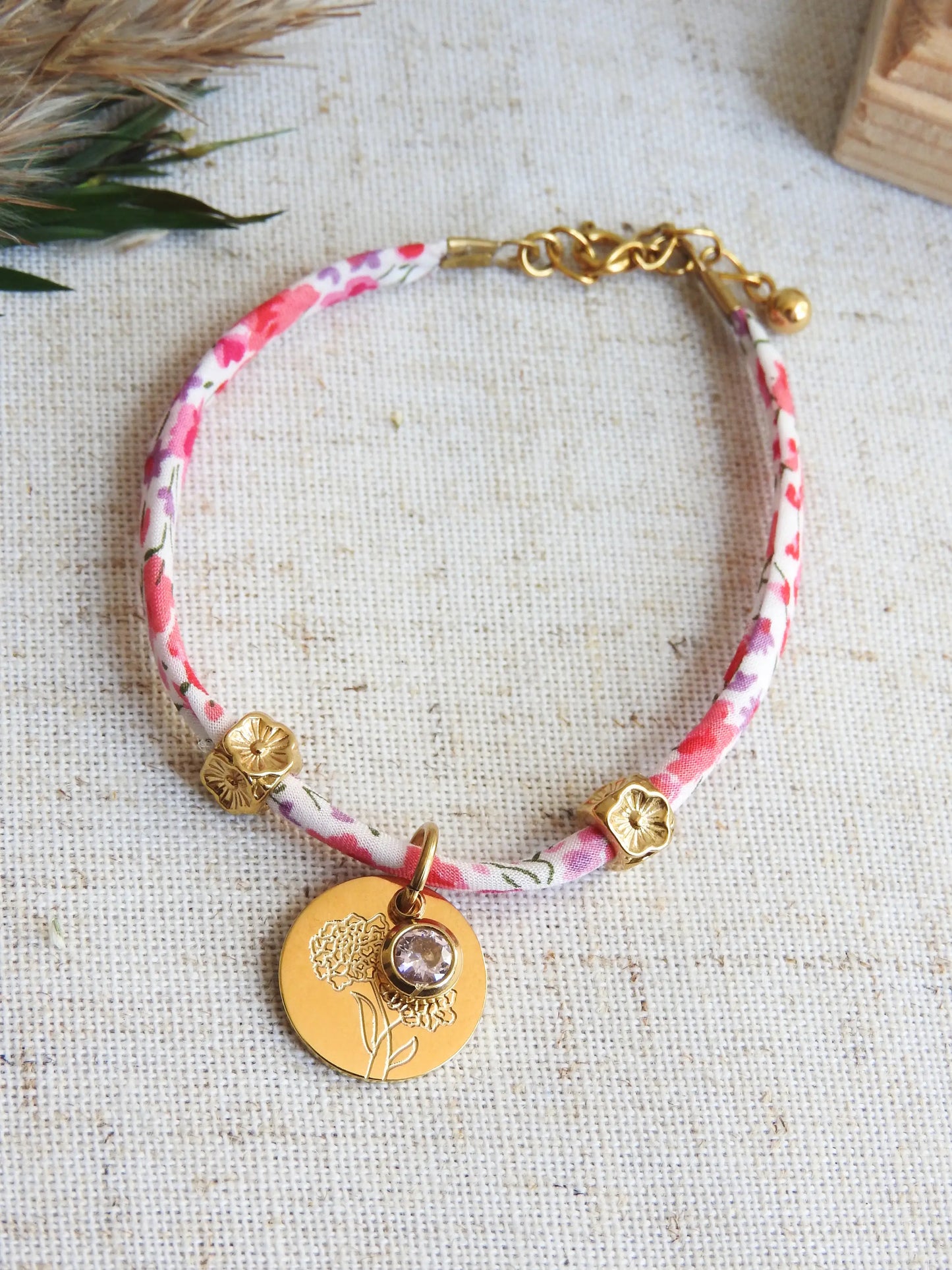Bracelet cordon liberty Phoebe aux couleurs rose et blanc floral. Pendentif Fleur Souci et pierre quartz rose de naissance en acier inoxydable doré. Bijou présenté à plat sur une nappe beige en coton.