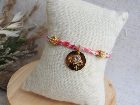 Bracelet cordon liberty Phoebe aux couleurs rose et blanc floral. Pendentif Fleur et pierre de naissance en acier inoxydable doré. Bijou présenté sur un mini-coussin beige en coton.