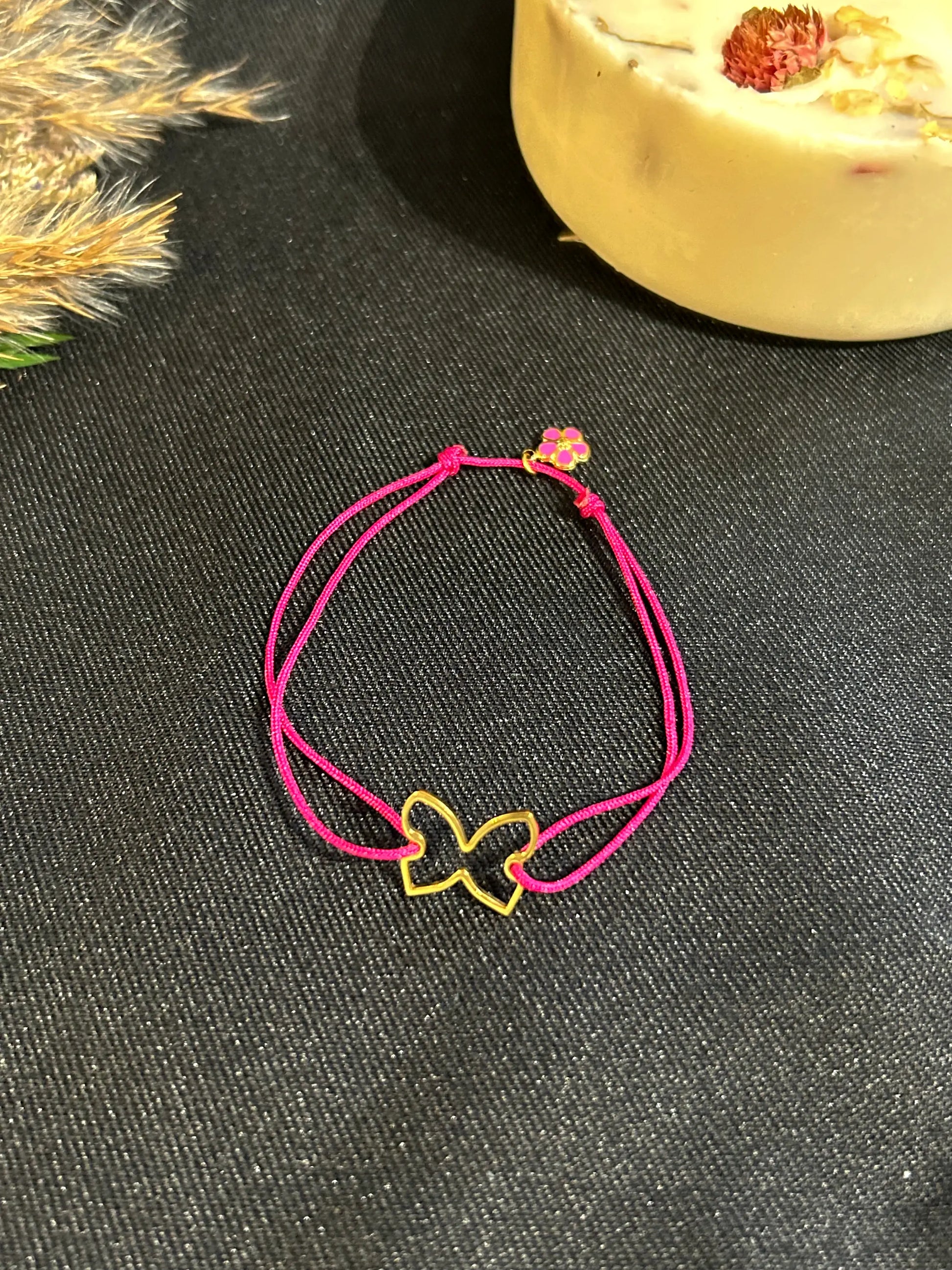 bracelet cordon en nylon tressé de couleur fuchsia, orné d'un papillon en acier inoxydable doré et une mini-breloque fleur fuchsia en acier inoxydable doré. Bijou posé sur une nappe noire décorée d'une bougie florale.
