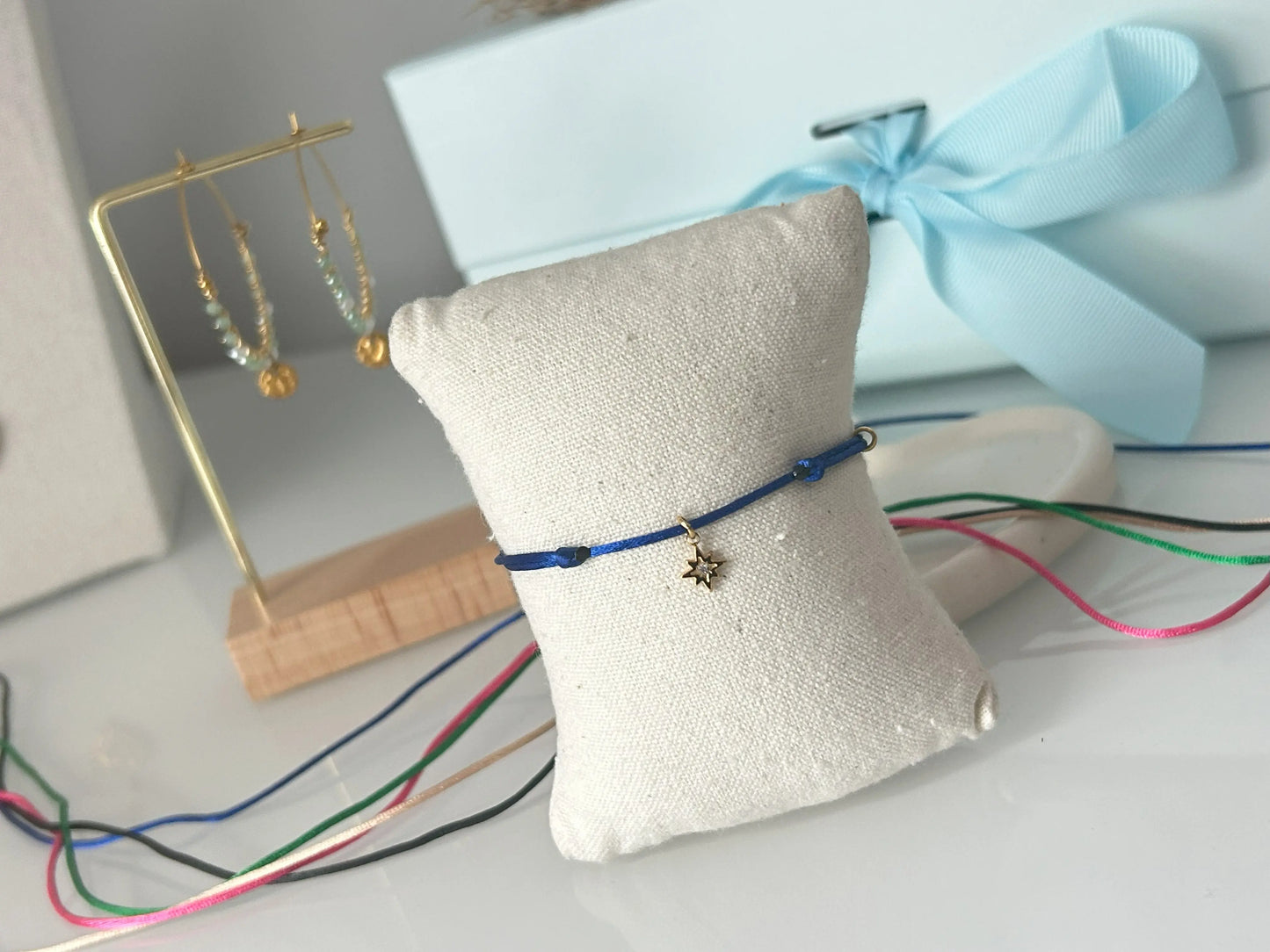 bracelets cordon ajustable en nylon bleu roi ornées d'une pampille étoile du nord avec une pierre en oxyde de zirconium et en acier inoxydable doré. Bijou présenté sur un mini-coussin beige en coton, posé sur une table décorée de cordon en nylon aux couleurs noir, vert électrique, champagne, rose fuchsia et bleu roi.