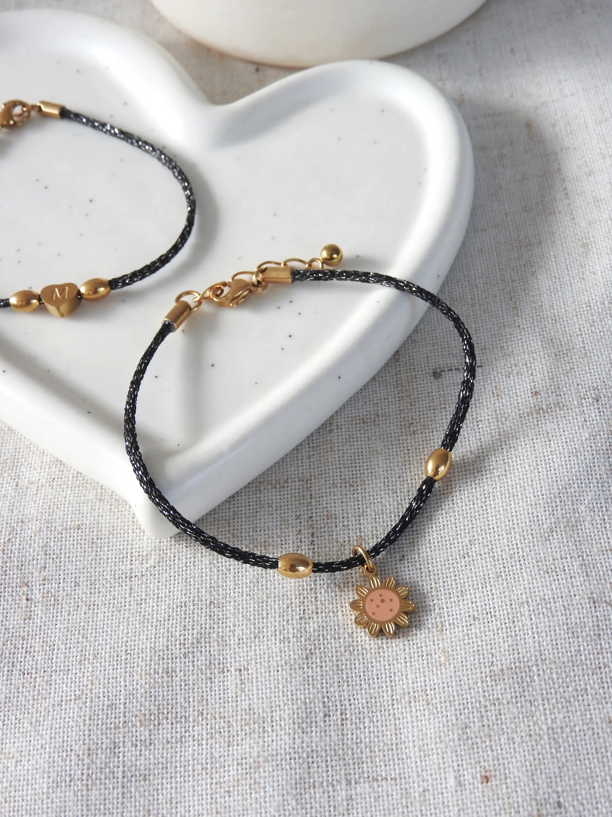 bracelet cordon scintillant mokuba du Japon de couleur noire orné d'une breloque tournesol couleur saumon en acier inoxydable et résine époxy. Bijou posé à plat sur une nappe beige et coupelle en céramique blanche.