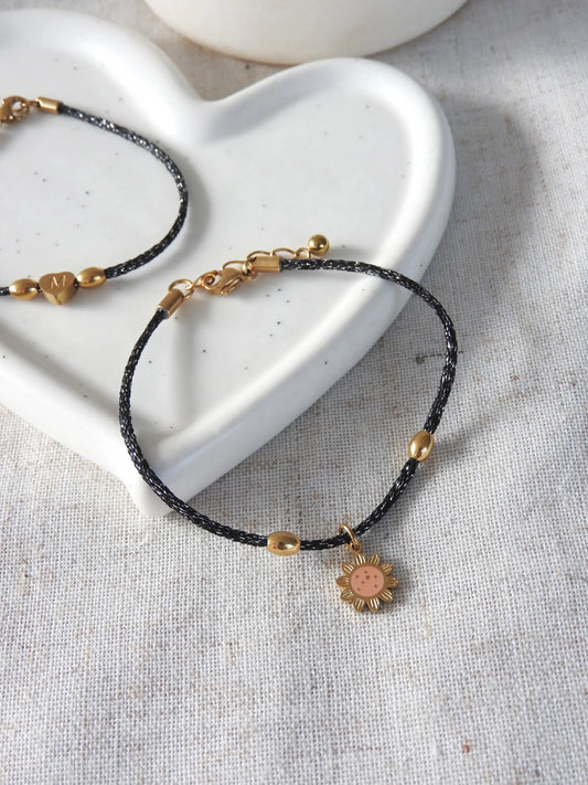 bracelet cordon scintillant mokuba du Japon de couleur noire orné d'une breloque tournesol couleur saumon en acier inoxydable et résine époxy. Bijou posé à plat sur une nappe beige et coupelle en céramique blanche.