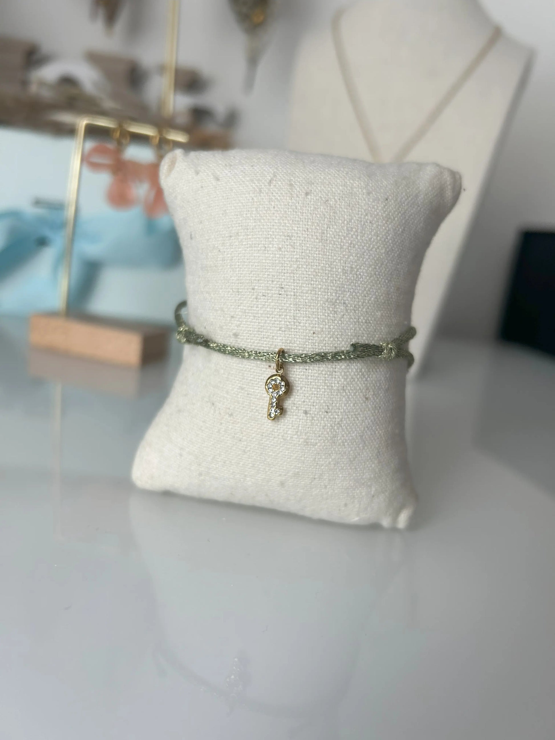 bracelet cordon scintillant vert olive à fil doré présenté sur un mini-coussin beige en coton.  Bijou orné d'une clé avec strass en acier inoxydable doré. En fond un buste avec un collier doré et une paire de boucles d'oreilles dorées et saumon sur un présentoir en bambou.