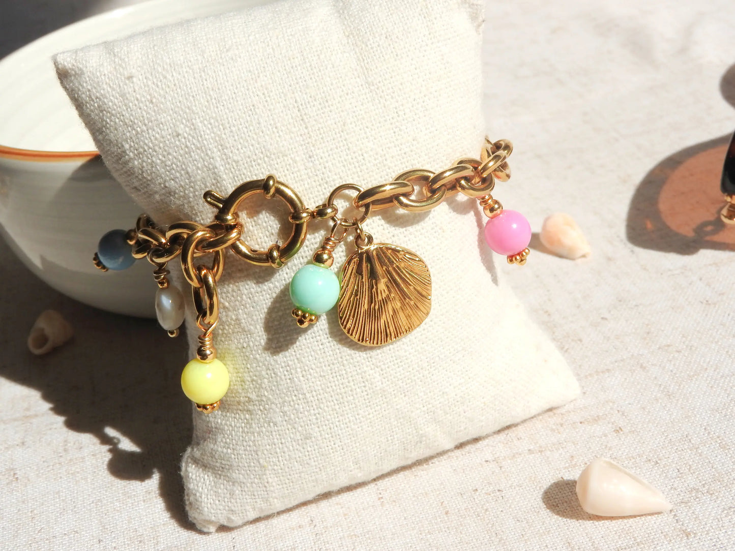 Bracelet larges mailles à breloques en acier inoxydable doré et perles colorées pastelles en acrylique posé sur un mini-coussin beige.