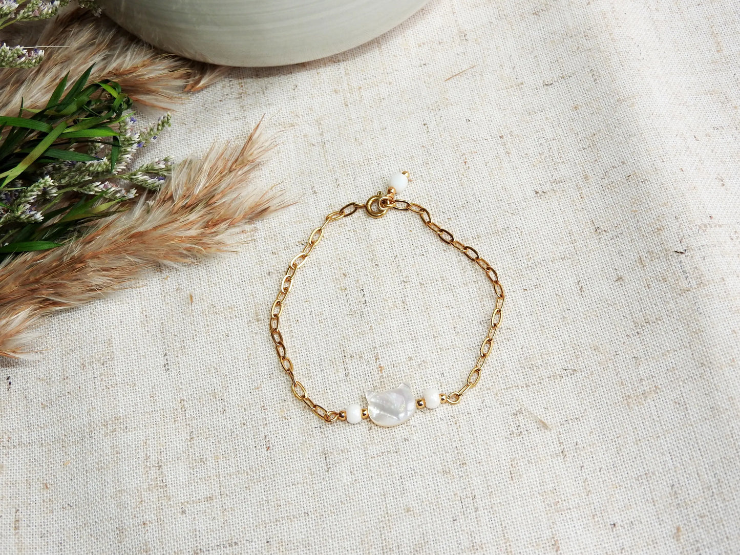 bracelet chaîne maille rectangle orné d'une perle chat en nacre blanche. Bijou posé sur une nappe beige décorée d'un mini-bouquet de fleurs.