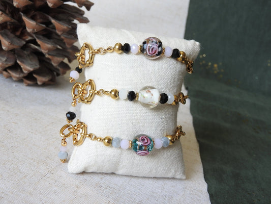 Trois bracelets mi-chaîne et mi-perles en verre, fermoir bouée en acier inoxydable doré. Bijoux présentés sur un mini-coussin beige en coton.