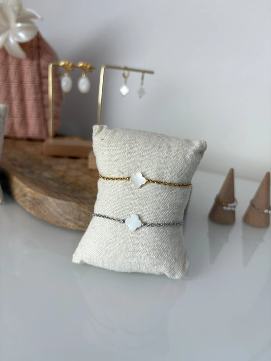 bracelets chaîne en acier inoxydable doré et argenté posés sur un mini-coussin en coton beige. Bijoux ornés d'une perle trèfle en nacre blanche. En fond, des bagues chaînette ornées de perles d'eau douce et un plateau en bambou avec des boucles d'oreilles présentées sur un présentoir métallique.
