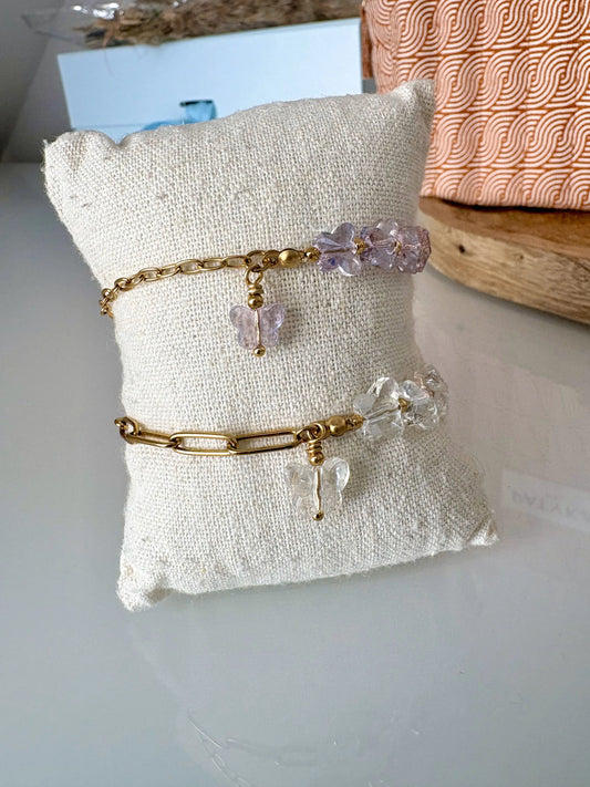 bracelets mi-chaîne en acier inoxydable doré et mi-perles de verre de forme papillon de couleurs mauve et blanche. Bijoux présentés sur un mini-coussin.