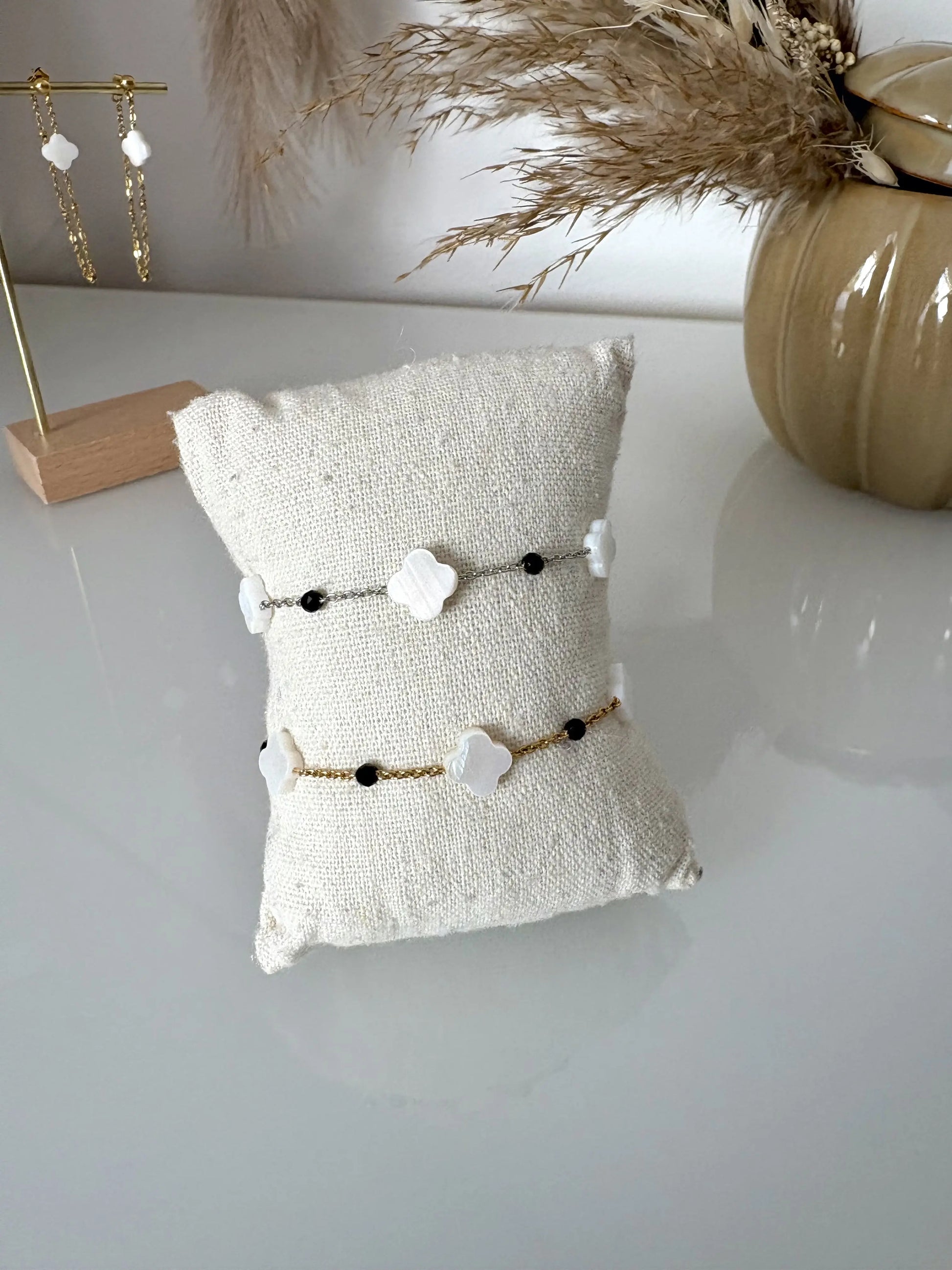 deux bracelets chaînes en acier inoxydable doré et argenté perlés de perles noires et trèfle en nacre blanche. Bijoux présentés sur un mini-coussin beige en coton posé sur une table décorée de brindilles, un pot en céramique kaki et des boucles d'oreilles pendantes.
