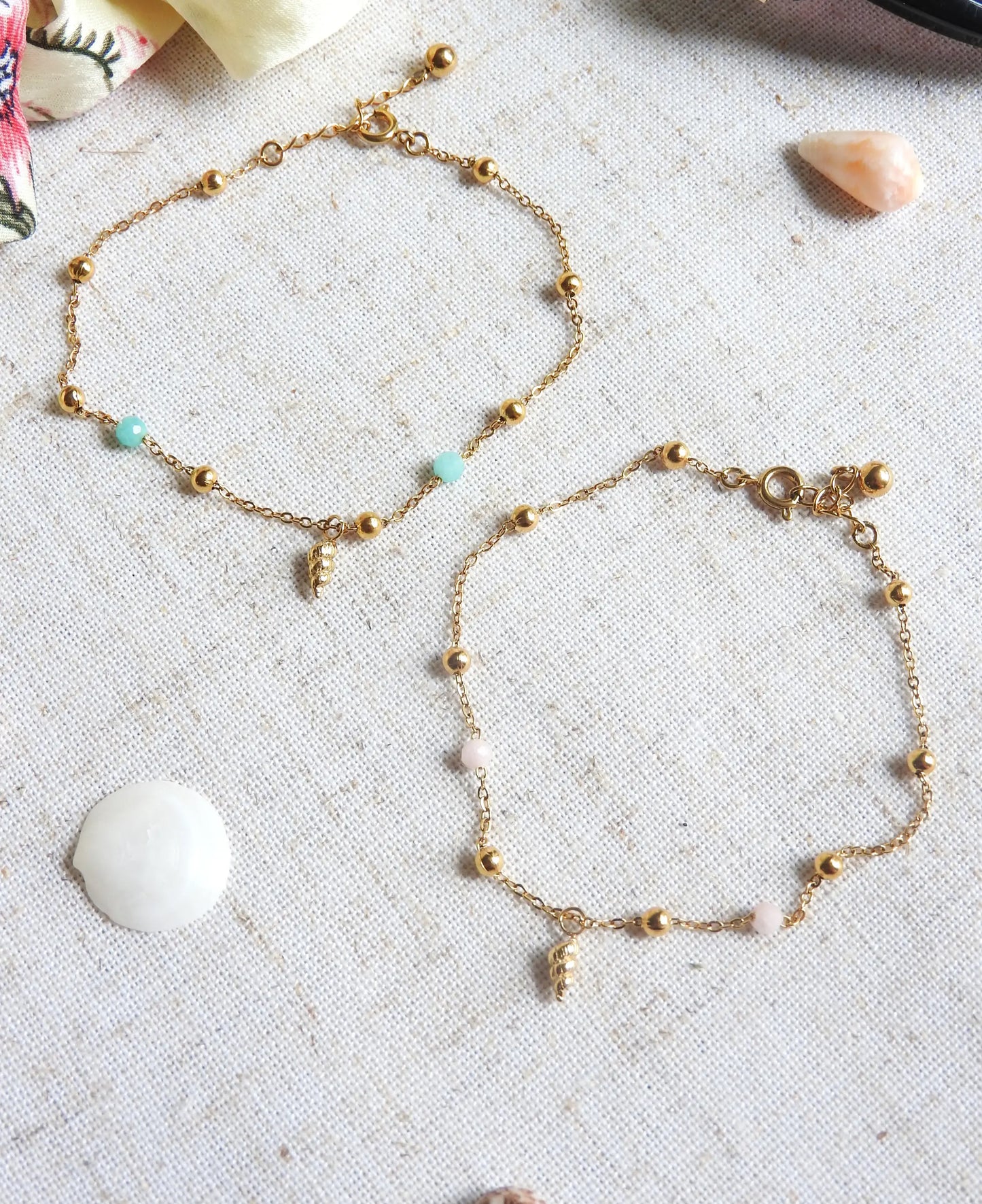 bracelets chaîne perlées en verre et acier inoxydable doré posés à plat sur une nappe beige décorée de coquillage de mer. Bijoux ornés d'une mini-breloque en coquillage doré.