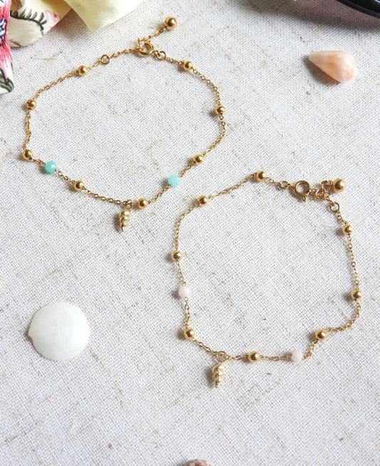 bracelets chaîne perlées en verre et acier inoxydable doré posés à plat sur une nappe beige décorée de coquillage de mer. Bijoux ornés d'une mini-breloque en coquillage doré.