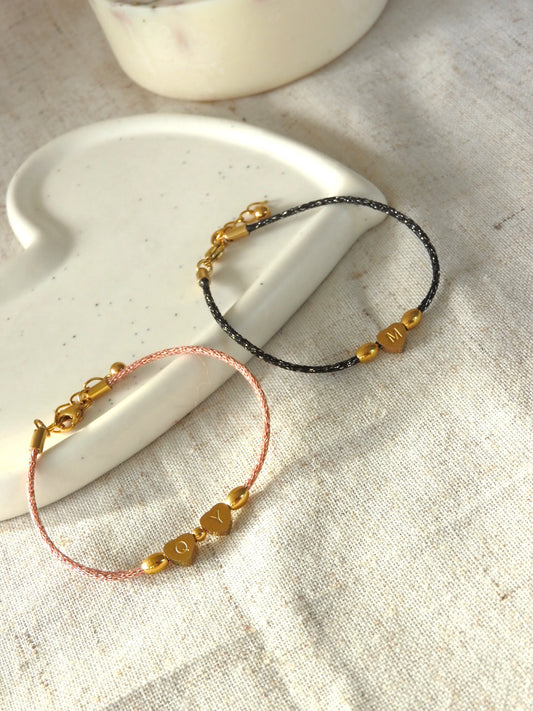 Bracelets cordon noir et saumon scintillant ornés de 3 perles dont une en forme de coeur plat gravée de la lettre M pour le bracelet noir et les lettres Q et Y pour le bracelet saumon en acier inoxydable doré. Bijoux posés sur une coupelle blanche en céramique.