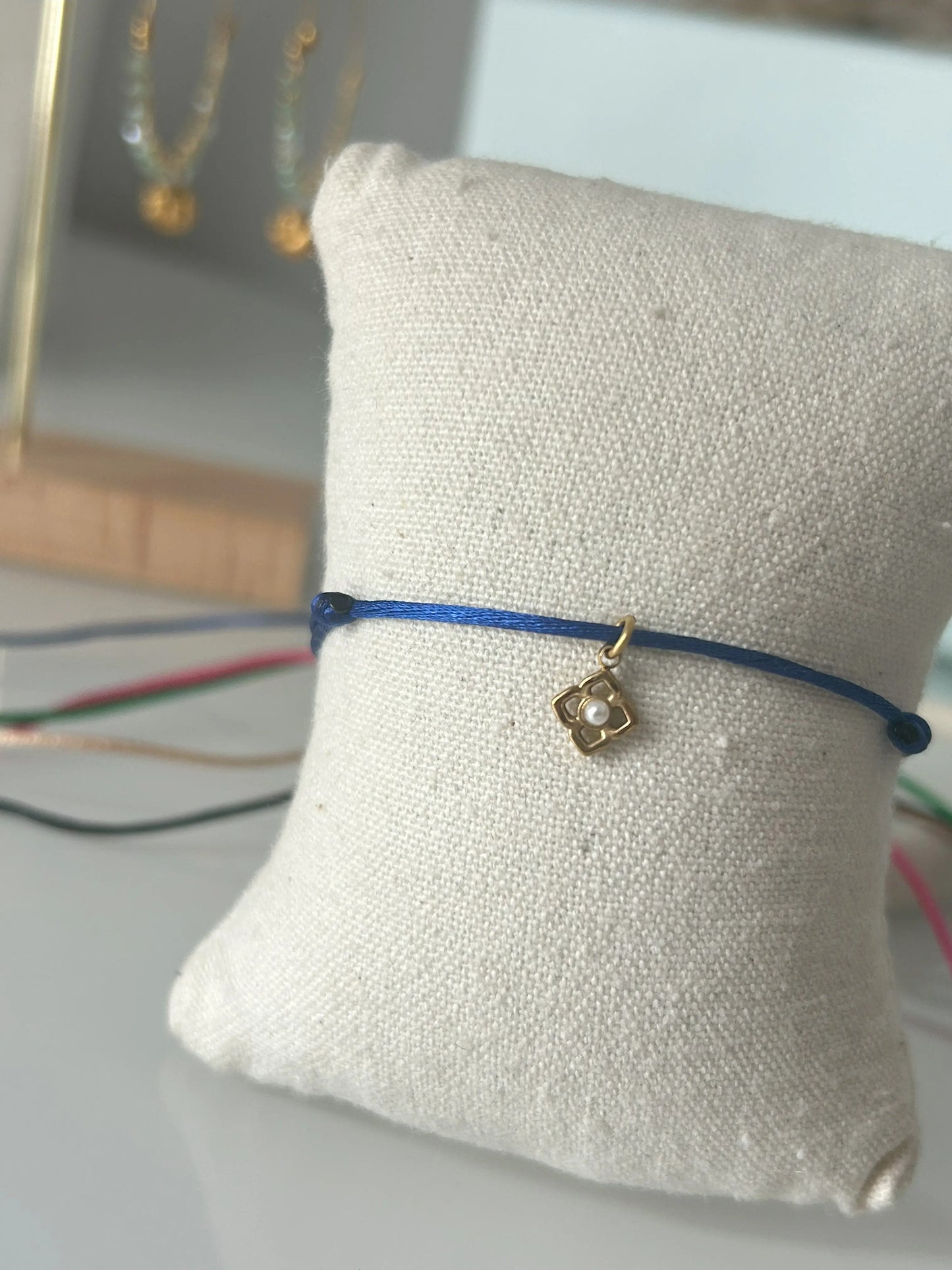 bracelet cordon ajustable en nylon bleu roi orné d'une pampille fleur avec une perle blanche imitation perle d'eau douce et en acier inoxydable doré. Bijou présenté sur un mini-coussin beige en coton.